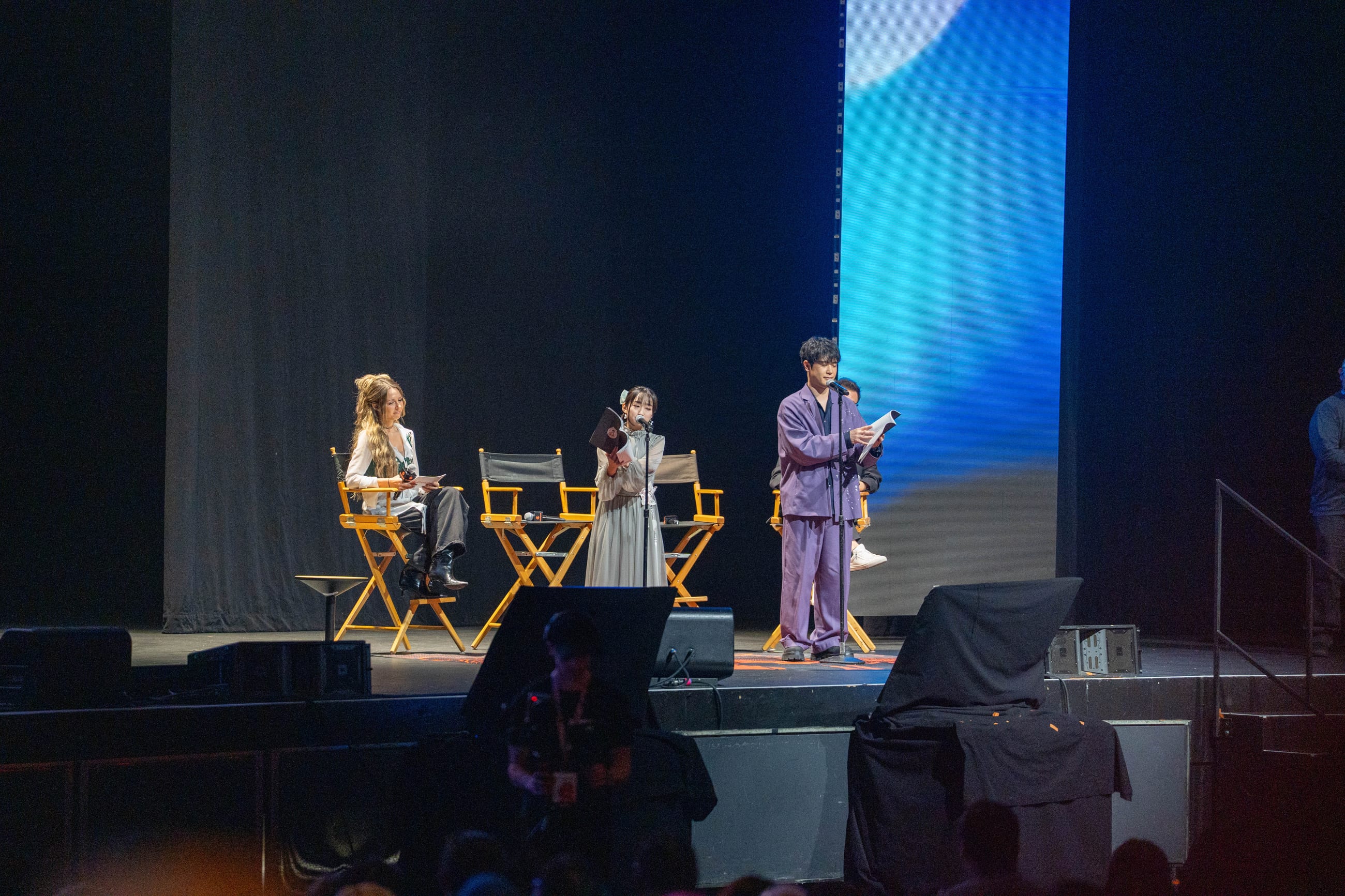 Panel in progress on the main stage, four speakers in director chairs against a lit blue screen
