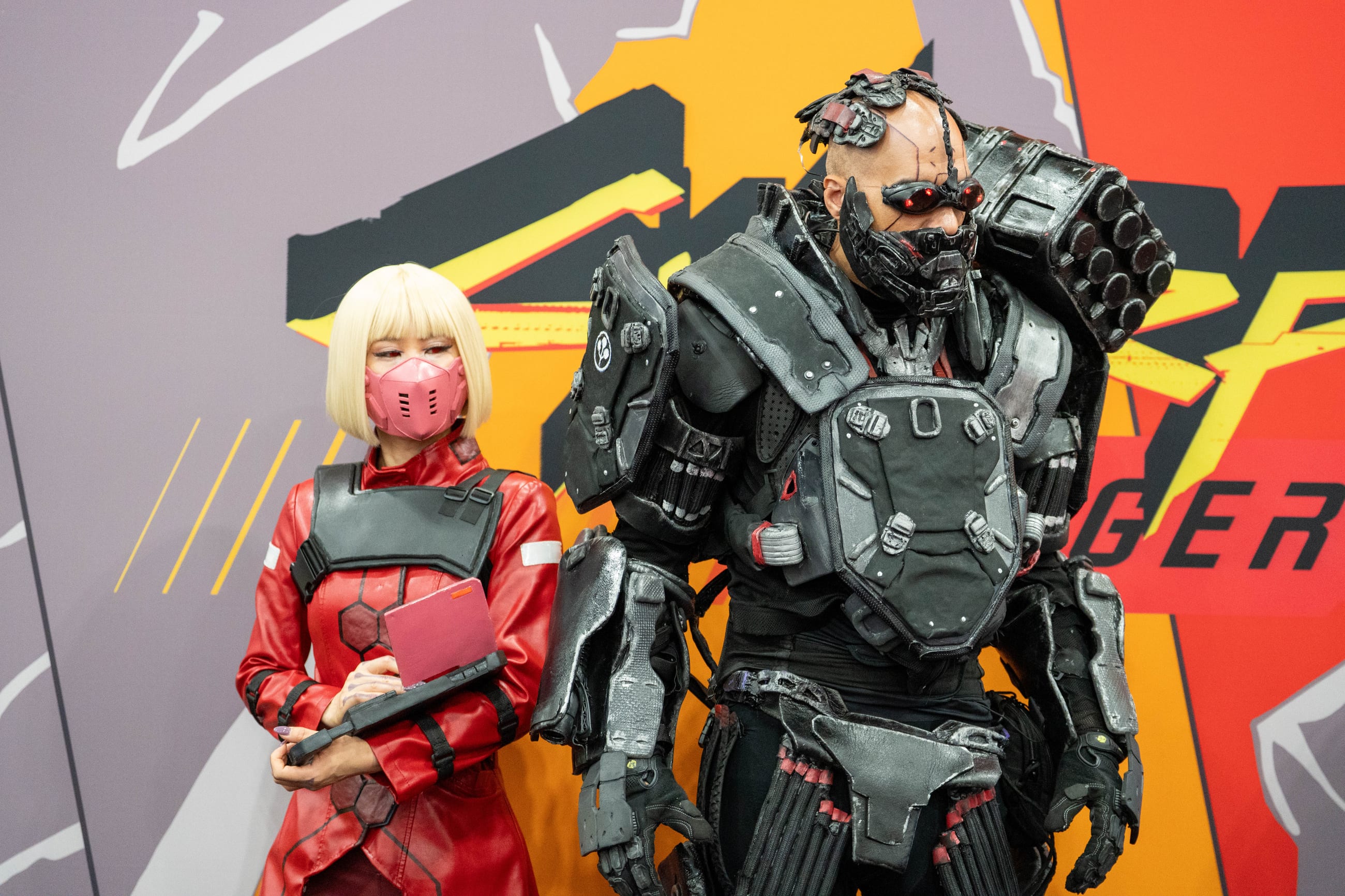 Two cosplayers in red and armored gray against a graphic orange and yellow activation wall
