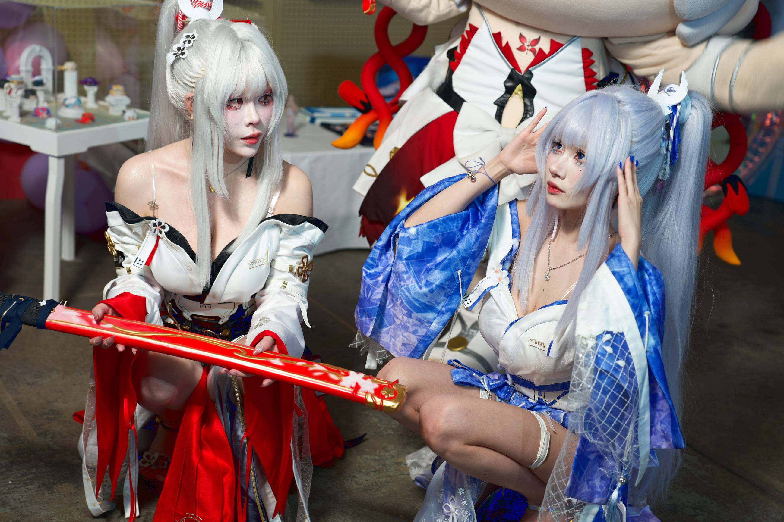 Two cosplayers kneeling side by side in white, blue and red anniversary outfits, holding a red blade between them