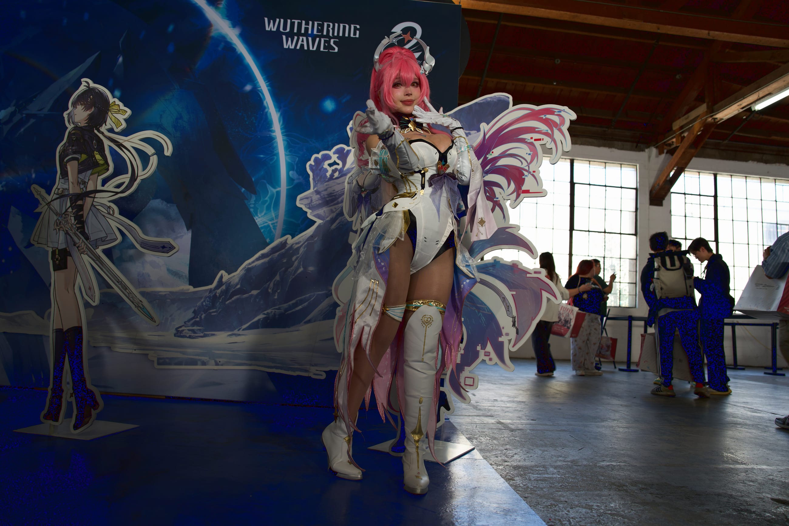 Cosplayer with pink hair and layered feathered wings standing before a key-art backdrop in a daylit hall